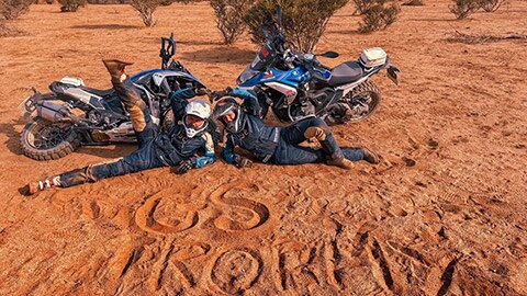 Obe vodičky medzinárodného ženského tímu ležia pred motocyklami v piesku, do ktorého napísali GS Trophy. 